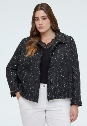 Žena oblečená v černé bundě s knoflíky a bílými puntíky přes černý top s krajkou a bílé kalhoty, stojící s jednou rukou v kapse.