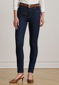 Temno modre skinny kavbojke z oranžnimi šivi, z zadnjim zadrgo in zanko za pas; v kombinaciji s smešnim slingback obutvijo v rjavi barvi.