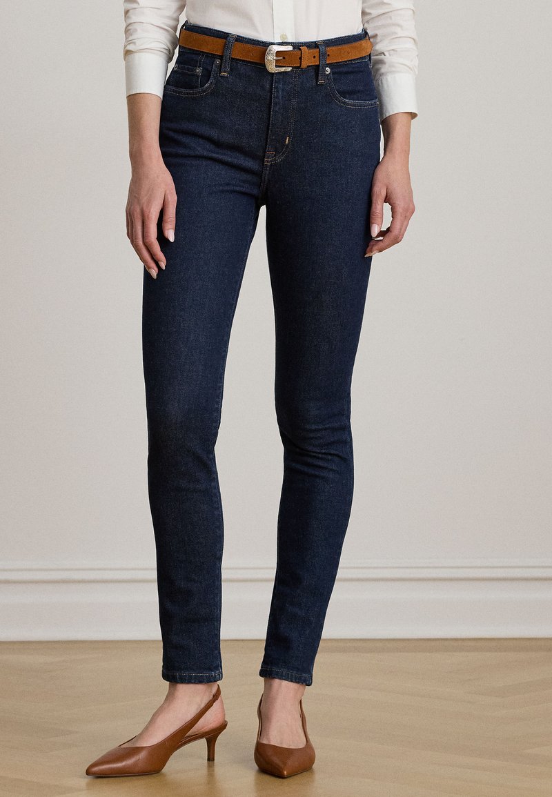 Temno modre skinny kavbojke z oranžnimi šivi, z zadnjim zadrgo in zanko za pas; v kombinaciji s smešnim slingback obutvijo v rjavi barvi.