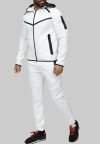 Survêtement athlétique blanc avec des accents noirs, comprenant une veste zippée avec une capuche et des poches zippées, associée à un pantalon fuselé assorti.