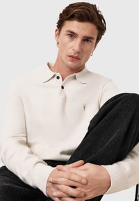 Pull polo tricoté couleur crème avec un col, deux boutons foncés et un petit logo brodé sur la poitrine. Porté avec un pantalon foncé.