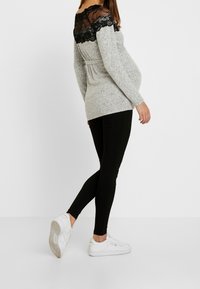 Haut gris à manches longues avec accents en dentelle noire aux épaules, associé à des leggings noirs et des baskets blanches. Coupe près du corps avec taille peplum.