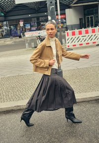 Veste courte en daim marron, col roulé blanc, jupe longue plissée grise et bottines à talons hauts pointues noires, marchant sur un pavement texturé.