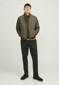 Ung mand står med hænderne i lommerne, iført en olivengrøn quilted jakke, mørk sweater, sorte bukser og sorte sko.