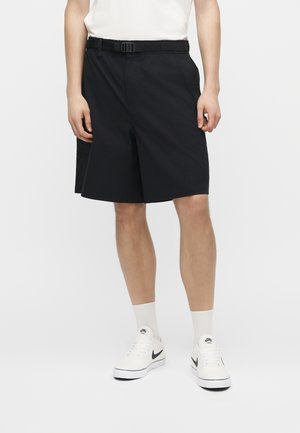 Άνδρας που φοράει μαύρα cargo σορτς μήκους μέχρι το γόνατο με ζώνη, λευκές κάλτσες και λευκά αθλητικά Nike με μαύρα λογότυπα Swoosh.