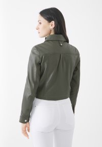 Olivgrüne, taillierte Lederjacke mit Hemdkragen, Druckknopfverschluss und Rückenschlitzdetail, kombiniert mit weißen Jeans.