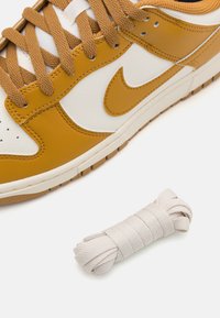 Nike sneakers med en gul læderoverdel, hvid læderside, brune snørebånd og en lysfarvet rund snor på siden.
