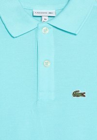 Błękitna koszulka polo z dwoma guzikami, metką Lacoste wewnątrz kołnierza oraz zielonym logo krokodyla na piersi.