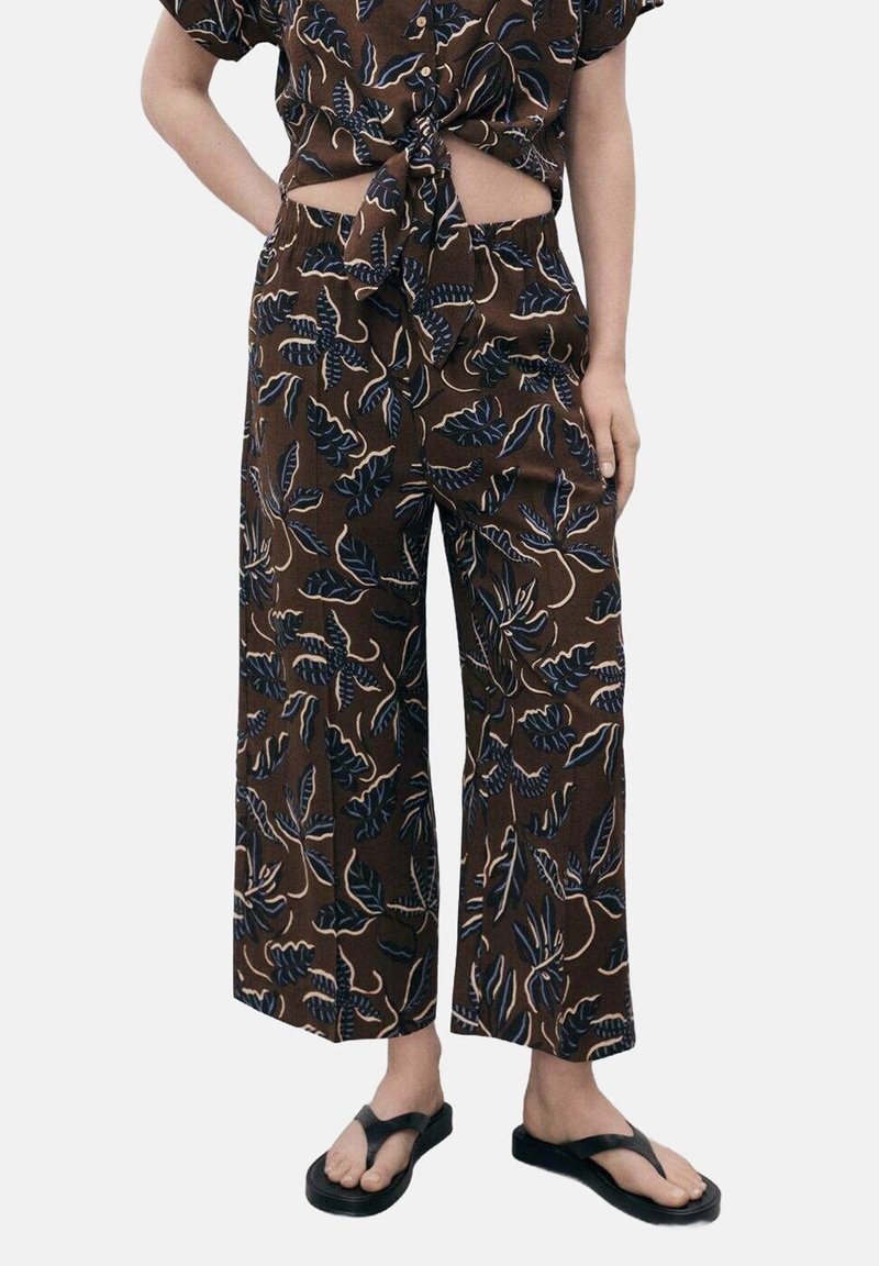 Pantalon large marron avec des motifs de feuilles bleu foncé, associé à un crop top noué assorti et des sandales noires type tongs, porté par une personne debout.