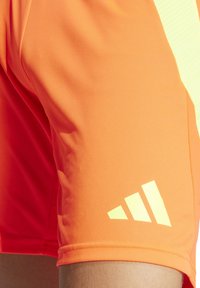 Oranje sportshorts van textuurstof, met een gele drie-stripes logo dicht bij de zoom en een contrasterend licht patroon aan de zijkant.