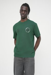 Πράσινο βαμβακερό t-shirt με κοντά μανίκια και στρογγυλή λαιμόκοψη, με το λογότυπο "TOMMY" και χειρόγραφες λεπτομέρειες στο μπροστινό μέρος.