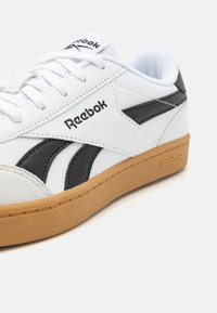 Balta ādas kedas ar melniem akcentiem un pelēku gumijas zoli. Reebok logo redzams sānos un uz mēlītes. Gluda tekstūra.