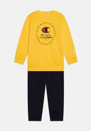 Felpa gialla con logo e testo neri, abbinata a pantaloni della tuta blu navy coordinati. Materiale in misto cotone, design casual, polsini a coste.