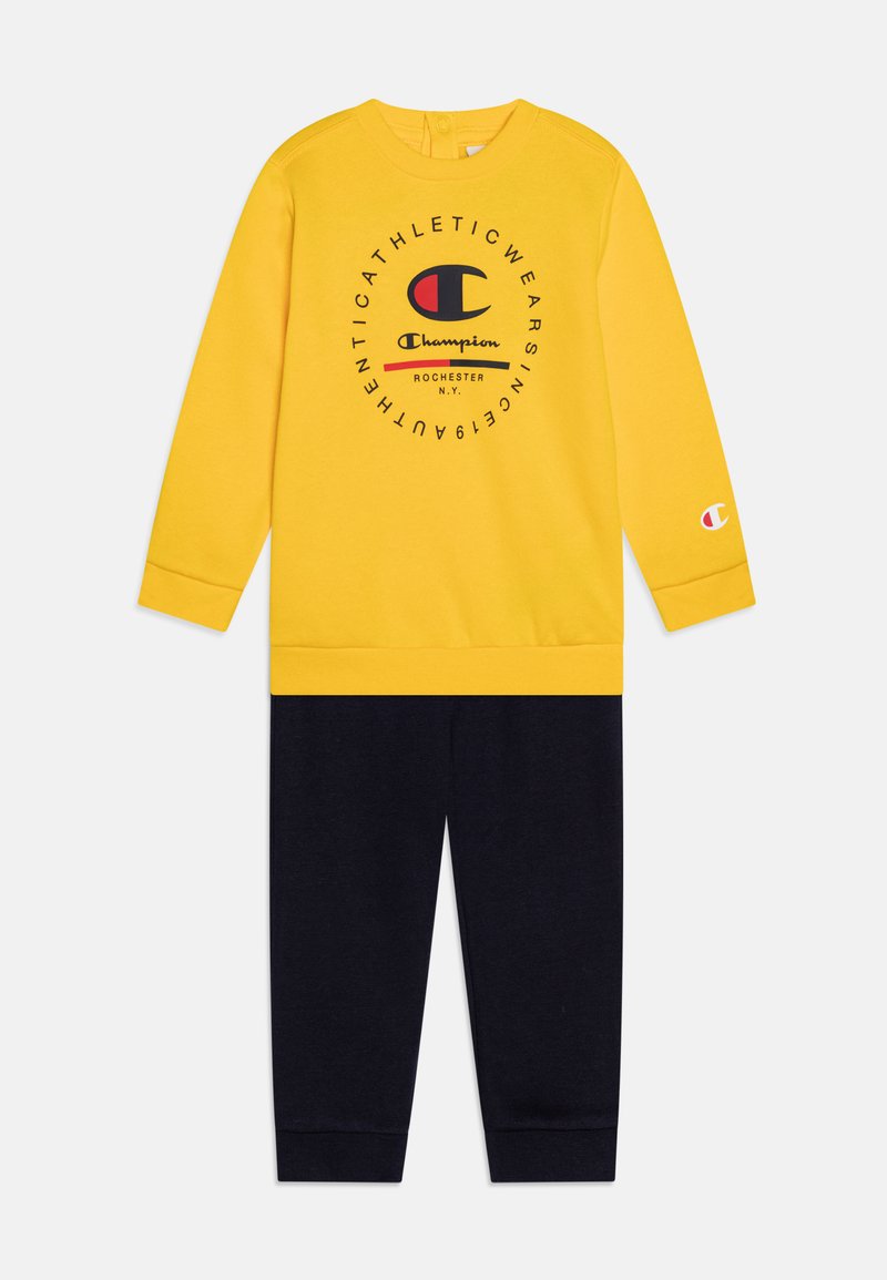 Sudadera amarilla con logo y texto en negro, combinada con pantalones deportivos azul marino a juego. Material de mezcla de algodón, diseño informal, puños acanalados.