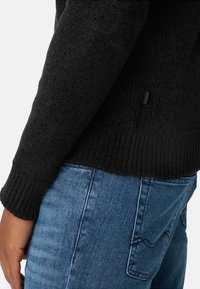 Zwarte gebreide trui met geribde boorden en zoom, gecombineerd met blauwe jeans met een zakdetail. De textuur is dik en gestructureerd.