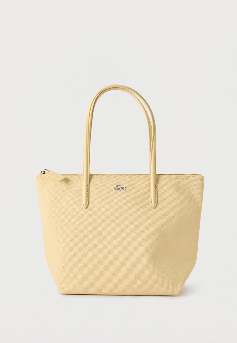 Sac fourre-tout jaune clair avec double poignée et un petit logo crocodile argenté sur le devant, posé sur un fond blanc uni.