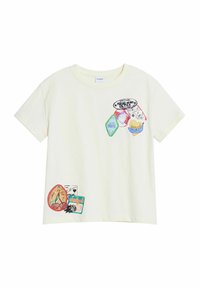 Camiseta de algodón de color beige claro con parches coloridos y bordados, que incluyen logotipos y texto, en varias formas y tamaños en la parte delantera.