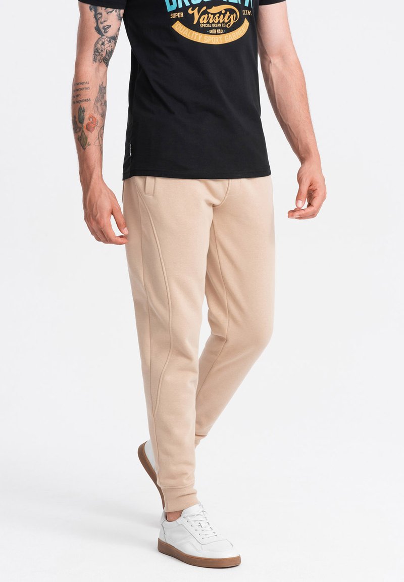 Beige joggings met voorkruiszakken, een gladde textuur, gesneden pijpen en geribbelde boorden, gecombineerd met witte schoenen en een zwart t-shirt.