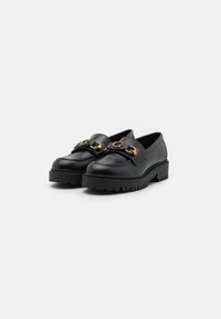 Mocassins en cuir noir avec une semelle noire épaisse, ornés d'un accent métallique doré prononcé sur le dessus. Texture lisse et design élégant.