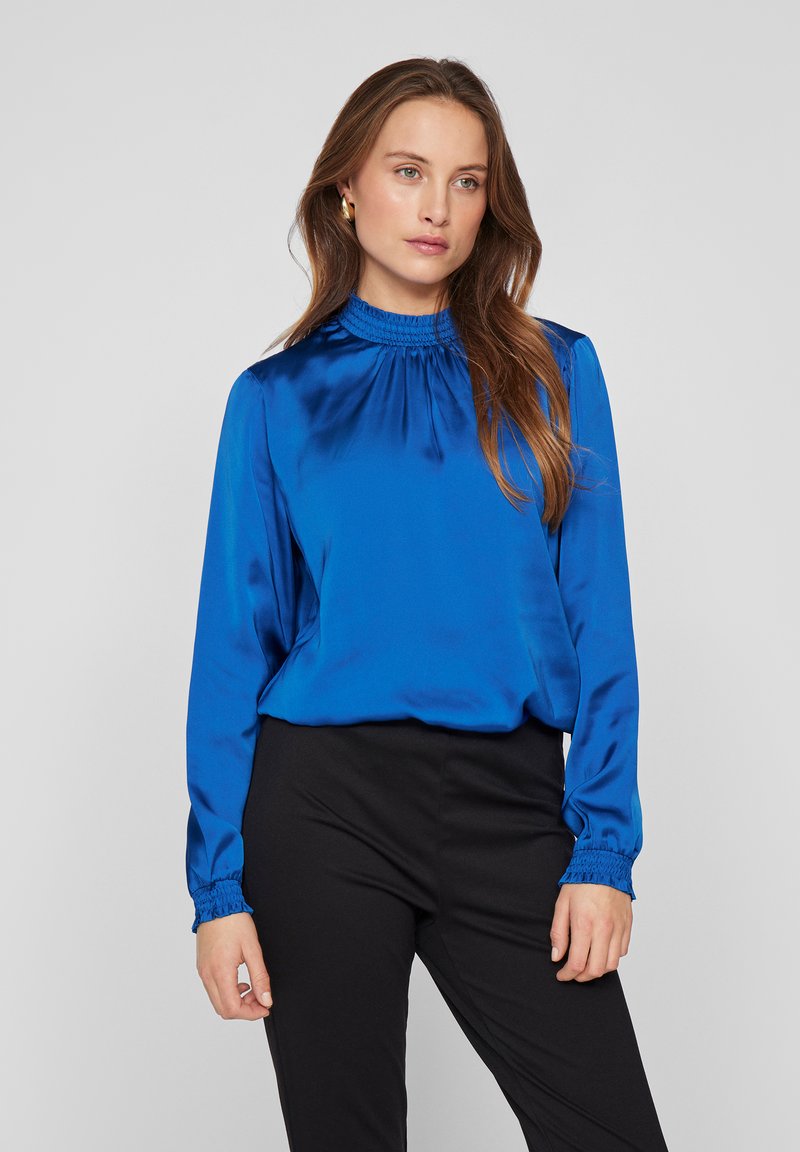 Helderblauwe satijnen blouse met lange mouwen, hoge elastische halslijn en elastische manchetten, gecombineerd met zwarte aansluitende broeken. Soepele textuur, ontspannen pasvorm.