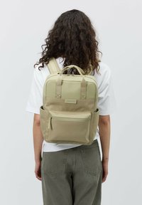 Beige ryggsäck med dubbla handtag på toppen och justerbara remmar, tillverkad av slitstarkt material. Har en framficka och sidoficka.