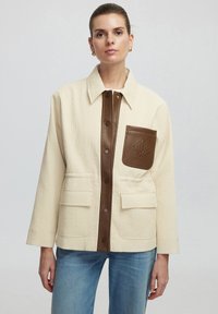 Gequilted crèmekleurige jas met bruin leren accenten, drukknoopjes en een borstzak; heeft een recht model en een gestructureerde stof.