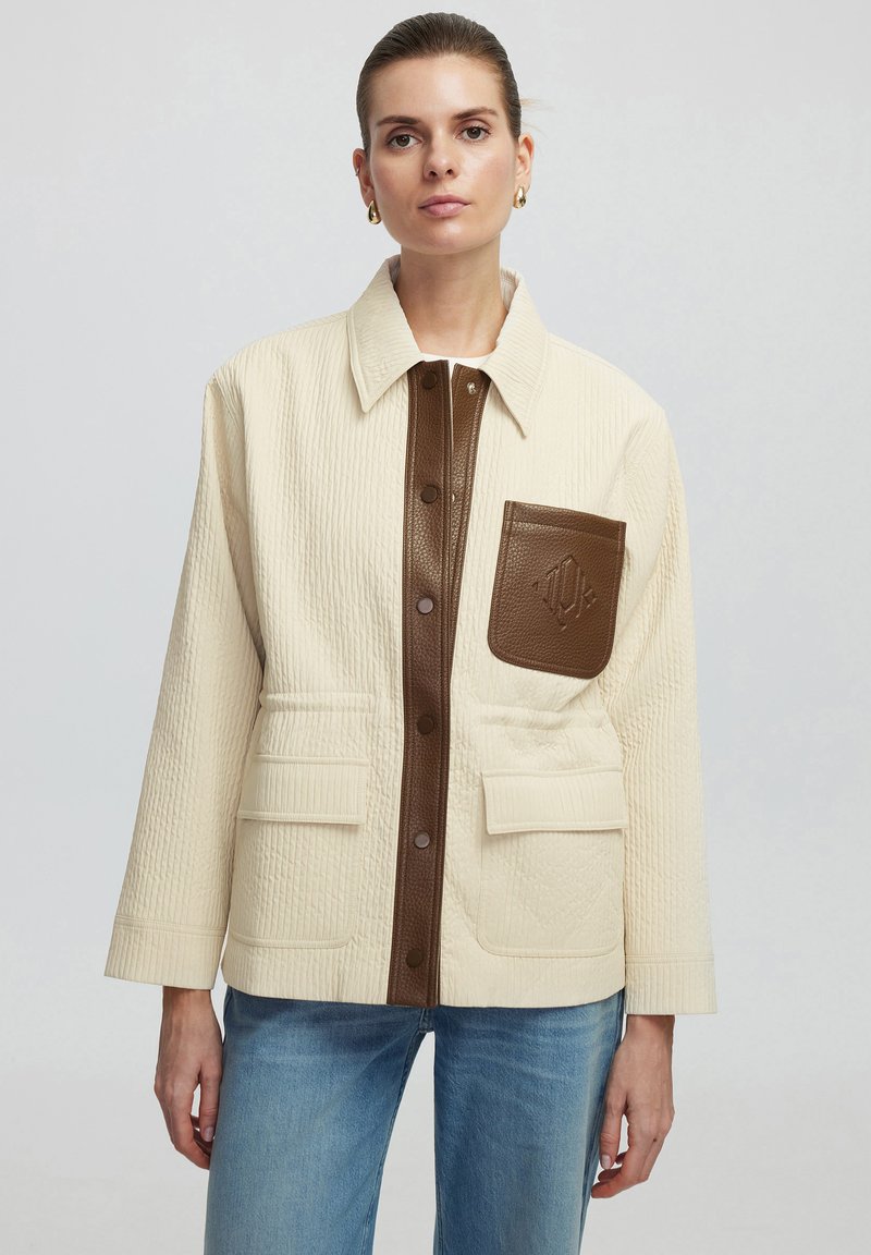 Gequilted crèmekleurige jas met bruin leren accenten, drukknoopjes en een borstzak; heeft een recht model en een gestructureerde stof.