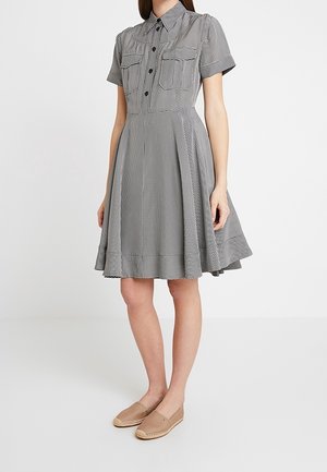 Femme portant une robe à carreaux noir et blanc avec des manches courtes, longueur genou, poches avant boutonnées, et des chaussures beige à enfiler, debout.