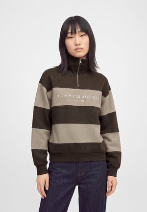 Jeune femme aux cheveux noirs et raides porte un sweat à capuche zippé rayé marron et beige Tommy Hilfiger et un jean bleu foncé.