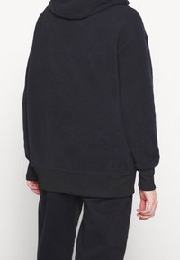 Černá fleecová mikina s texturovaným povrchem, kapucí a spadlými rameny. Obsahuje černé ribbané lemy a manžety pro těsné padnutí.