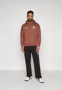 Άνδρας που στέκεται φορώντας ένα καφέ φούτερ με λογότυπα, μαύρο παντελόνι με λογότυπα και λευκά παπούτσια, μπροστά σε έναν απλό ανοιχτό γκρίζο φόντο.