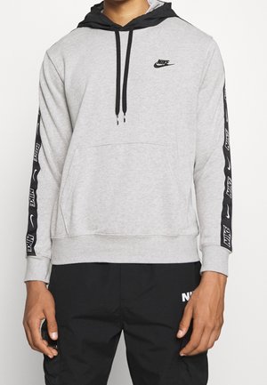 Mężczyzna noszący jasnoszarą bluzę z kapturem Nike z czarnym kapturem i paskami z logo na rękawach, w połączeniu z czarnymi spodniami Nike.