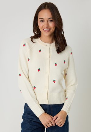 Vrouw die een crèmekleurige cardigan draagt met geborduurde aardbeien, glimlacht, armen ontspannen, tegen een effen lichte achtergrond.