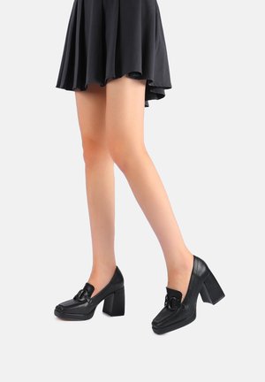 Scarpe nere in pelle con tacco a blocco e punta arrotondata, con un fiocco sulla parte anteriore e una superficie liscia. Abbinato a una gonna nera a ruota.