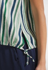 Widok z boku osoby ubrane w zielono-biało-niebieski paskowany top z bocznym rozcięciem i związanym dołem, w połączeniu z granatowymi spodniami.