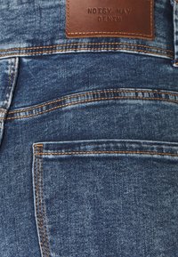 Vaqueros azules de mezclilla con un acabado texturizado, con costuras naranjas y una etiqueta de cuero marrón en la cintura que dice "NOISY MAY DENIM".