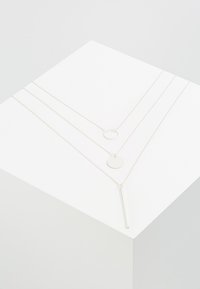 Silverfärgad lagerhalsband med tre distinkta kedjor: en tunn med en vertikal stav, en rund hänge och en cirkulär länk.