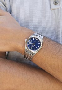 Silvrig rostfritt stål armbandsur med en blå texturerad urtavla, silverfärgade timmarkörer och datumsdisplay. Bärs på ett handled mot en grå bakgrund.