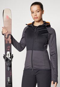Black and gray zip-up ski jacket with an orange hood accent, worn by a person holding a ski. Glossy ski with bindings attached on one side.