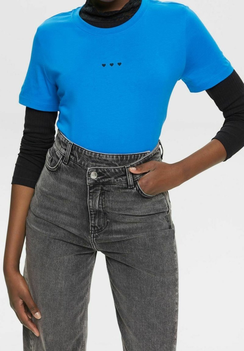 Lysblå kortærmet t-shirt med tre sorte hjertegrafikker på brystet, parret med højtaljede sorte denimjeans.