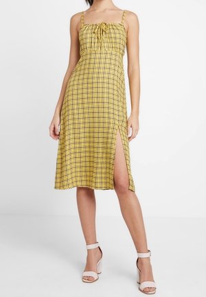 Vestido a cuadros amarillos con tirantes finos, cuerpo fruncido, abertura lateral y tejido ligero. Se lleva con tacones de tiras de color claro.