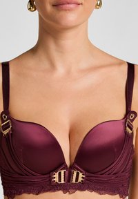 Soutien-gorge en satin bordeaux avec des bonnets plongeants, des bordures en dentelle, des éléments décoratifs en or et des bretelles ajustables. Texture lisse et design structuré.
