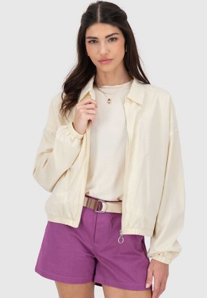 Lichtbeige vest met rits en een losse pasvorm, voorzien van elastische manchetten, gedragen over een crèmekleurige top en paarse korte broek met een beige riem.