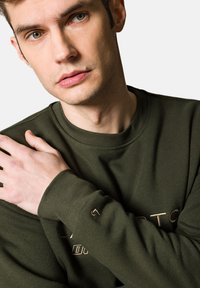 Ciemnozielony sweter z okrągłym dekoltem i ściągaczami. Złote napisy z przodu dodają kontrastu do materiału.