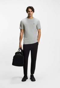 Szara koszulka z okrągłym dekoltem z małym logo, w połączeniu z czarnymi zwężającymi się spodniami i czarnymi sportowymi butami. Trzyma czarny plecak.