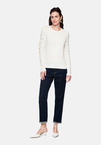 White long-sleeve top with gathered detail on sleeves, paired with dark blue cuffed jeans and white pointed-toe heels.
