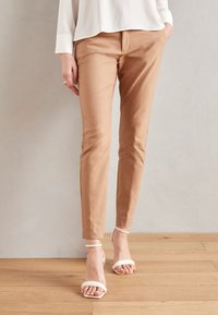 Žena v béžových slim-fit kalhotách a bílých sandálech na podpatku s pásky stojí na dřevěné podlaze před hladkou zdí.