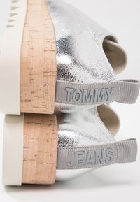 Chaussures métalliques argentées avec une semelle intermédiaire en liège, arborant des étiquettes en relief "TOMMY" et "JEANS" ainsi qu'une semelle extérieure en caoutchouc blanche. Design à bout rond.