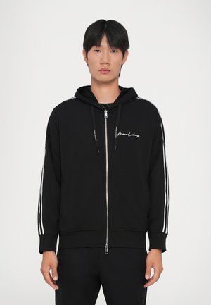 Mladic u crnoj dukserici s patentnim zatvaračem i bijelim trakama sa strane, s "Armani Exchange" logotipom na prsima, stoji nasuprot jednobojnoj pozadini.