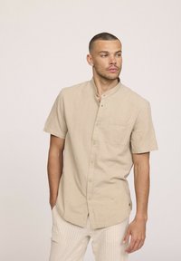 Chemise beige à manches courtes avec un col montant, une poche poitrine unique et un boutonnage sur le devant ; fabriquée en tissu léger et texturé.